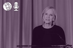[ai] A woman speaking at a podium in front of a curtain, engaged in a presentation. She has shoulder-length blonde hair and wears a black top. Icons of a globe and a microphone are in the upper left corner.